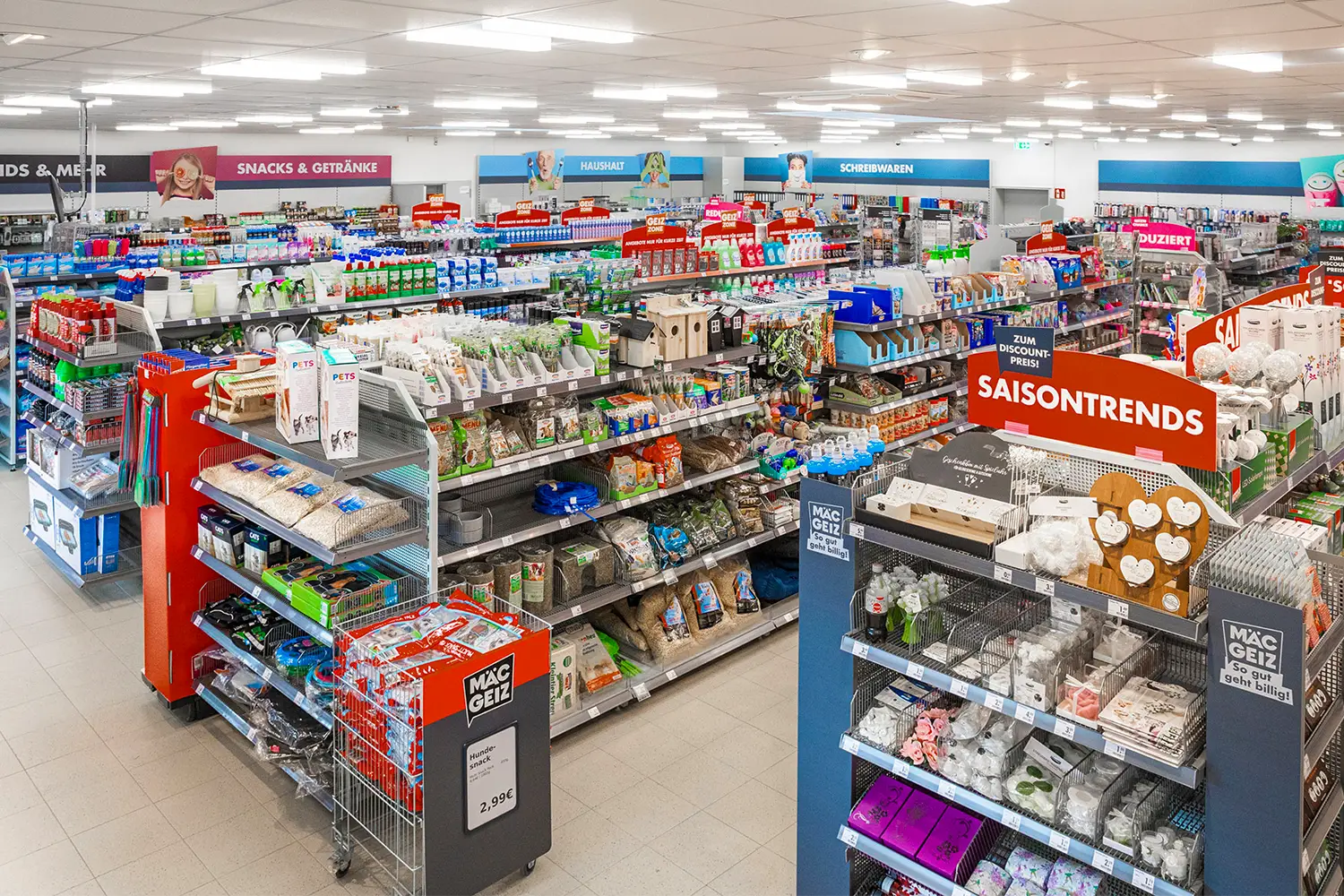 Innenansicht eines gut sortierten Discounters mit Regalen voller Haushaltswaren, Snacks, Schreibwaren und Tierbedarf, übersichtlich beschriftet mit Kategorie-Schildern.