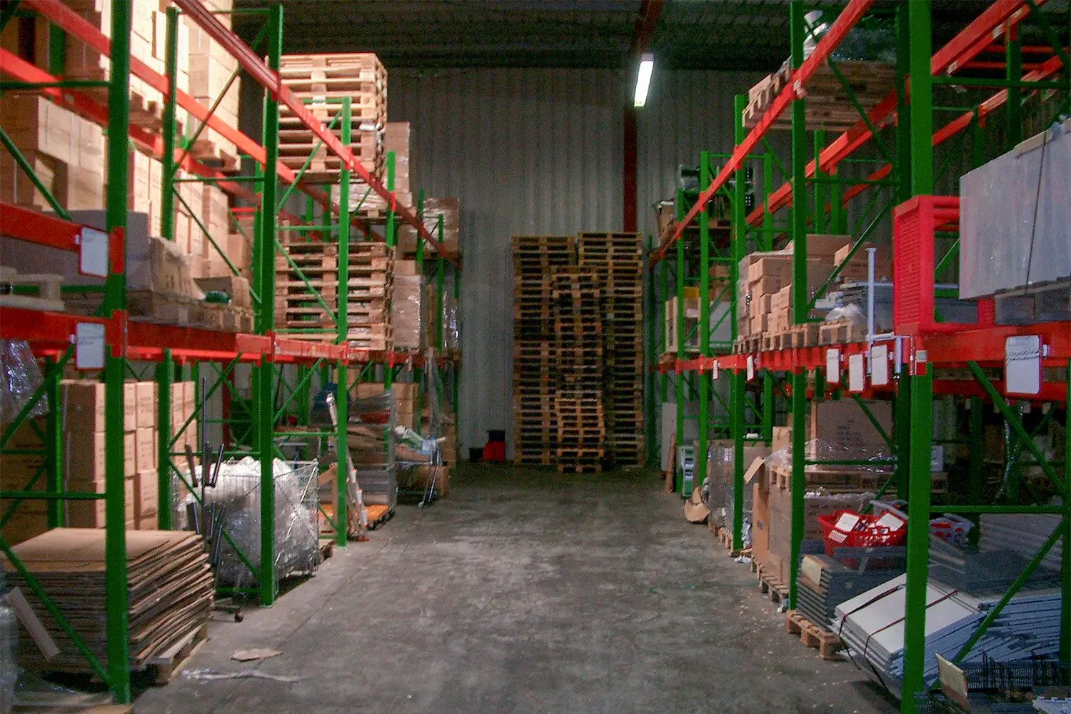 Blick in eine Lagerhalle mit hohen Regalen, gefüllt mit Kartons, Paletten und verschiedenen Materialien, die in einem schmalen Gang gestapelt sind.