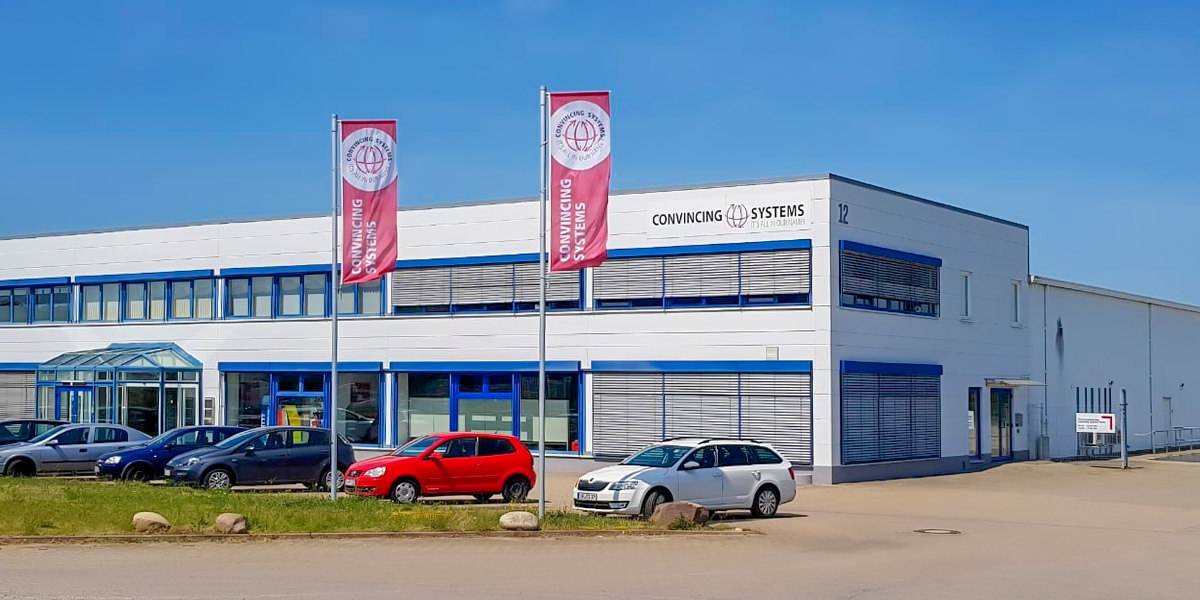 Beschreibung: Firmengebäude von Convincing Systems mit moderner Glasfassade und mehreren Fahrzeugen im Vordergrund.