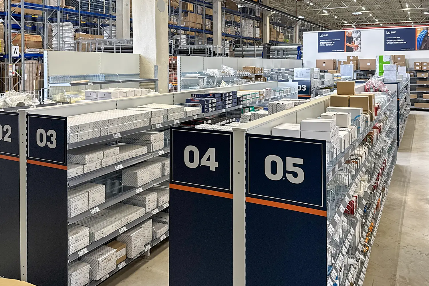Ein Großhandelsgeschäft mit mehreren Regalreihen, in denen zahlreiche kleine Produkte lagern; die Regale sind nummeriert (z. B. 04 und 05) und gut sortiert.
