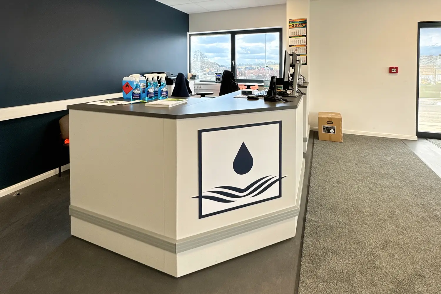 Ein moderner Empfangsbereich mit weißem Tresen, einem Logo mit Wassertropfen, Büroausstattung und Blick durch Fenster nach draußen; an der Wand hängt ein Kalender für das Jahr 2025.