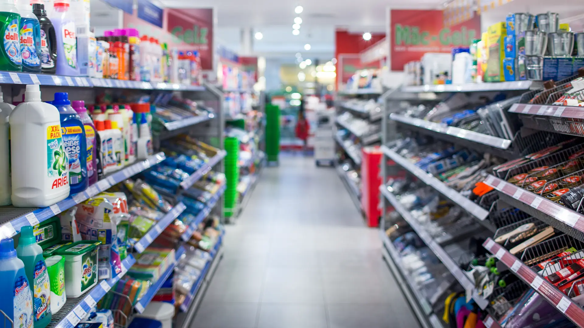 Ein langer Gang im Einzelhandel mit dicht bestückten Regalen, die zahlreiche Haushalts- und Kleinteile in Drahtkörben bereithalten. Die Gänge sind gut organisiert und übersichtlich.