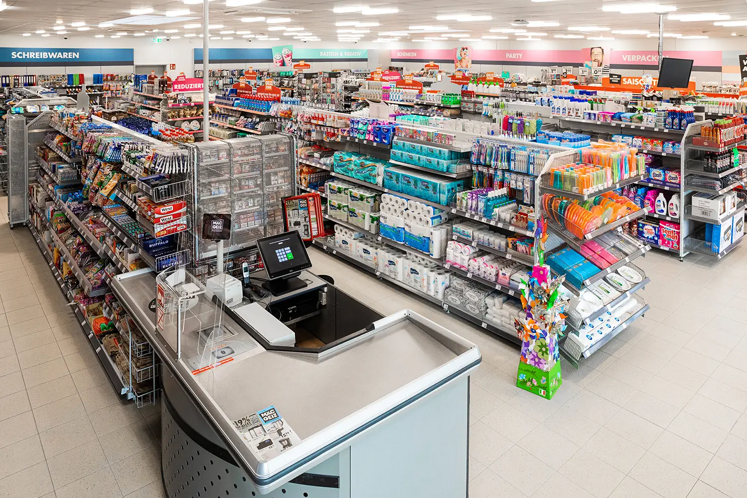 Heller Kassenbereich in einem Non-Food-Discounter mit gut sortierten Regalen, freundlicher Atmosphäre und übersichtlicher Warenpräsentation. Ideal für Einzelhandel, Ladeneinrichtung und Einkaufsambiente.