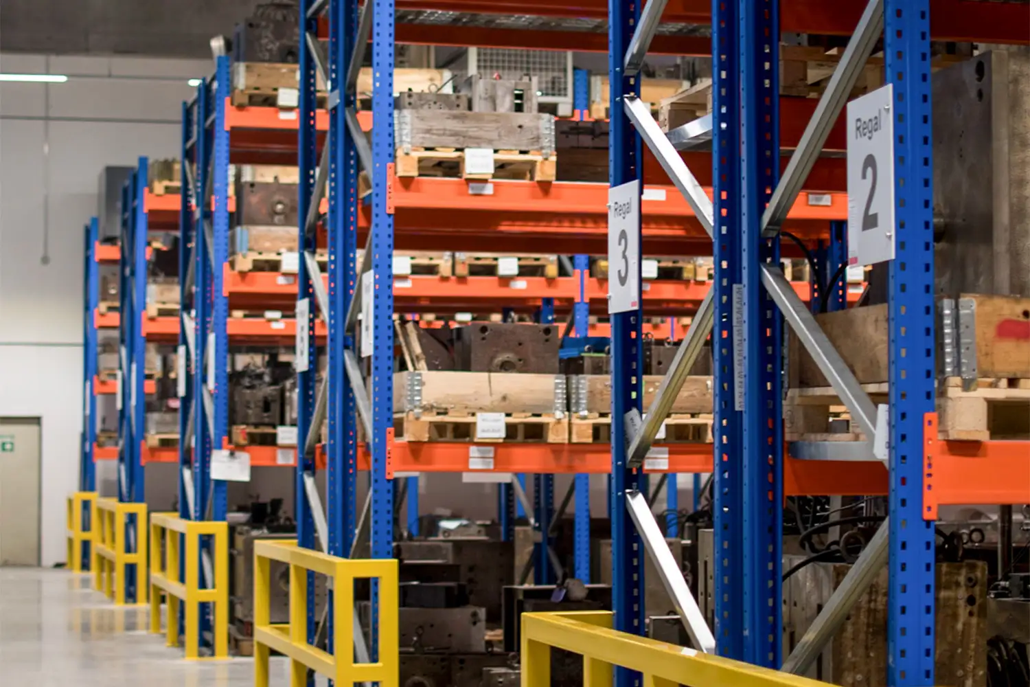 Ein Lagerraum mit Schwerlastregalen aus blauem Stahl und orangefarbenen Trägern, bestückt mit schweren metallischen Werkzeugformen auf Paletten; die Regale sind nummeriert (z. B. Regal 2 und 3) und durch gelbe Sicherheitsgeländer voneinander getrennt.