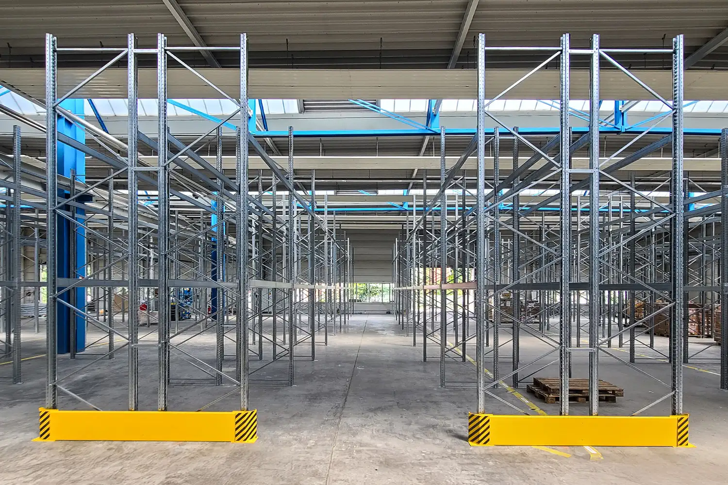 Ein großes Lager mit leeren, verzinkten Metallregalen in mehreren Reihen; im Vordergrund gelbe Schutzbarrieren und viel Tageslicht durch Oberlichter.