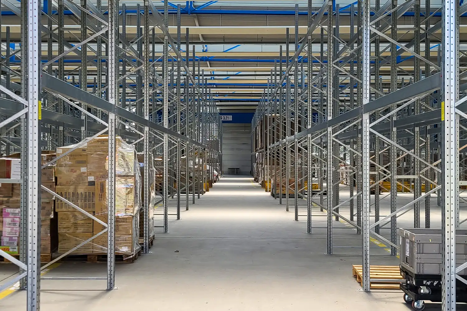 Lager mit Hochregalen, befüllt mit palettierten Kartons; mittig ein breiter Gang mit Blick auf ein geschlossenes Tor im Hintergrund; links und rechts verschiedene Ladeeinheiten.